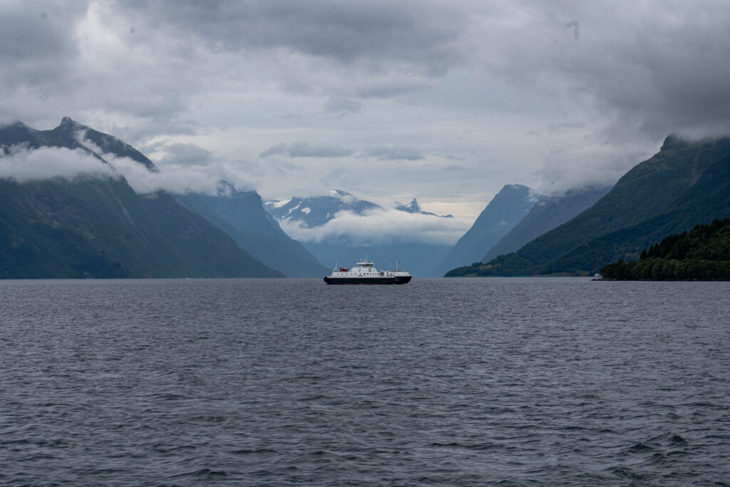 Photographie d'un paysage de Norvège montrante un bateau centré sur l'image au milieu d'un fjord entouré de montagne aves des nuages bas et une ambiance paisible