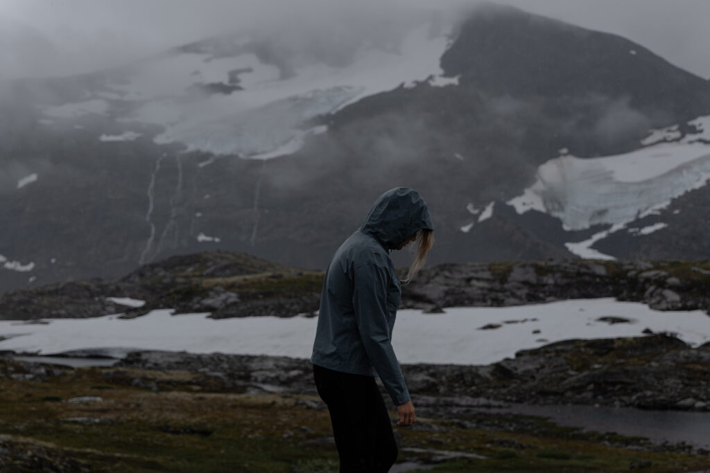 Manon LORY, photographe de profil portant une veste de pluie et une capuche avec un glacier de Norvège en arrière plan.