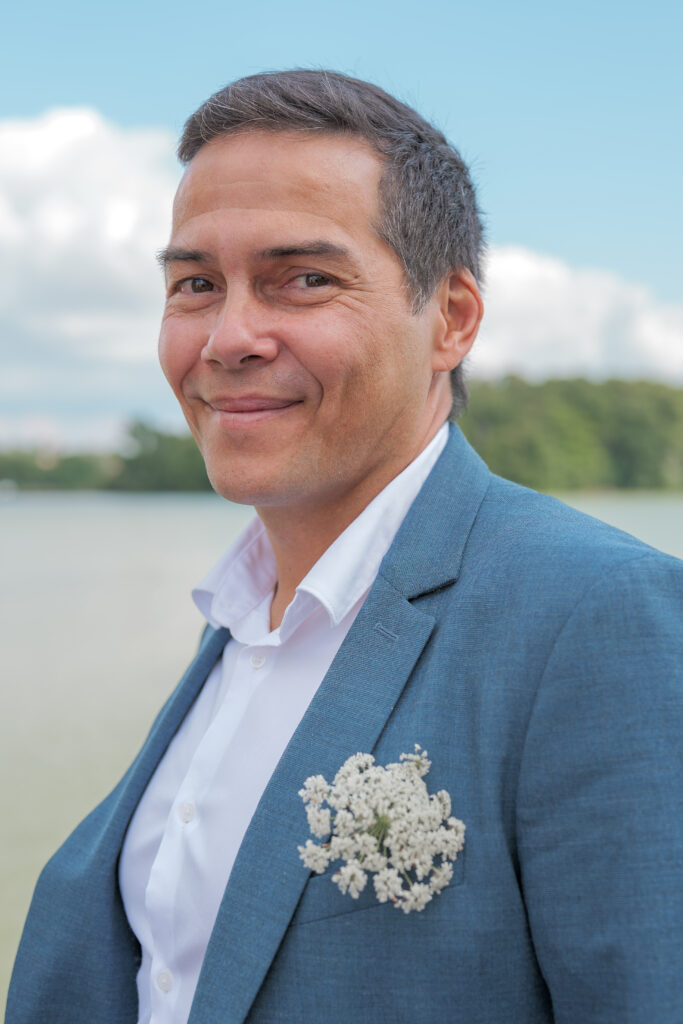 Invité de mariage portant une fleur dans la poche et souriant à l'objectif avec en arrière plan un étang  et des arbres au loin