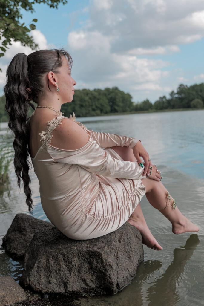 Portrait d'une jeune femme assise sur un rocher au bord de l'eau et habillé d'une jolie robe champagne regardant vers un étang