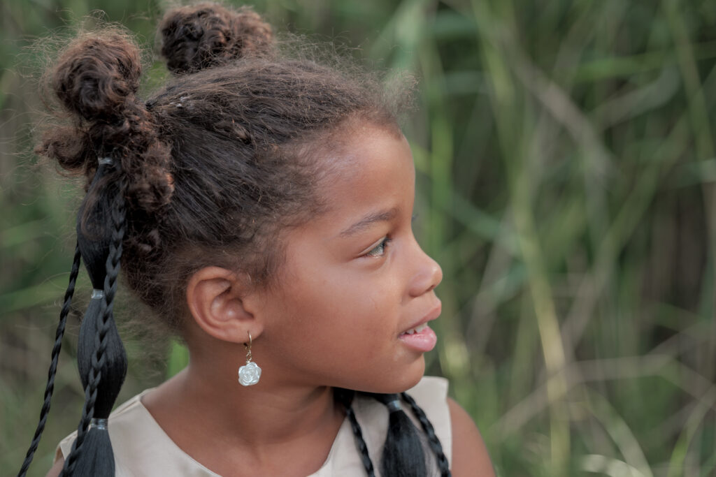 Portrait de profil d'une petite fille coiffée de deux petits chignons sur le dessus de la tête ainsi que de jolies tresses, regardant vers la droite et mettant en valeur sa boucle d'oreille