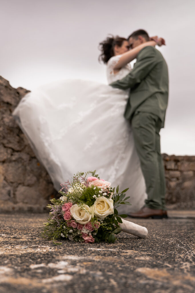 Premier plan focus sur le bouquet de la marié et flou d'arrière plan sur les mariés s'enlaçant, la robe de la mariée vole au vent