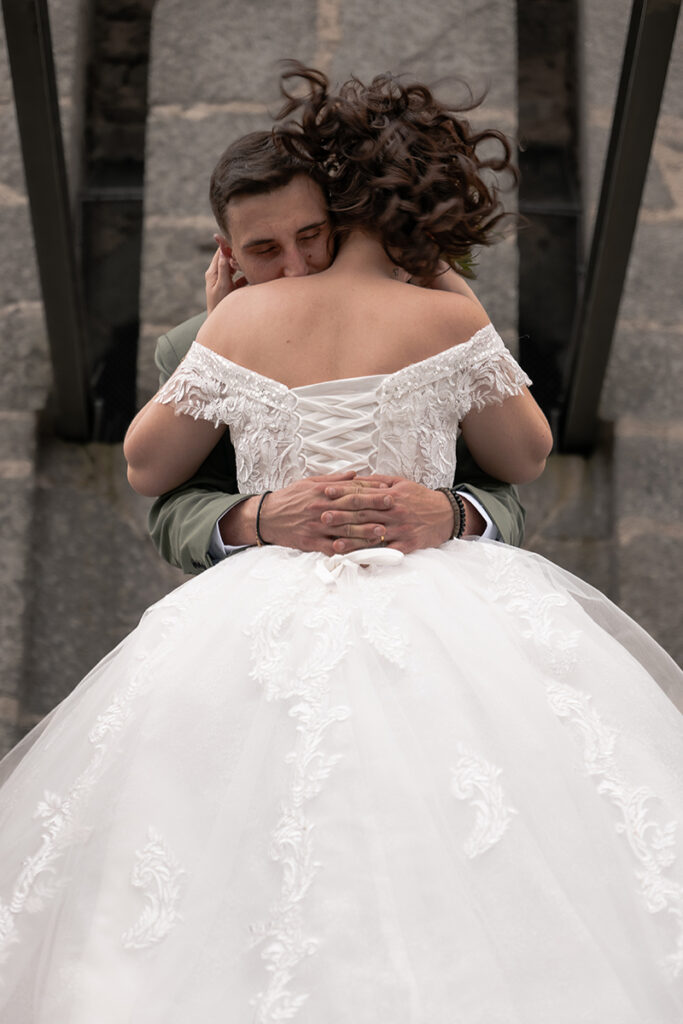 Photographie d'un couple de marié où l'on voit la mariée de dos et le mari l'enlaçant au dessus de la taille et fermant les yeux avec une tour médiévale en arrière plan
