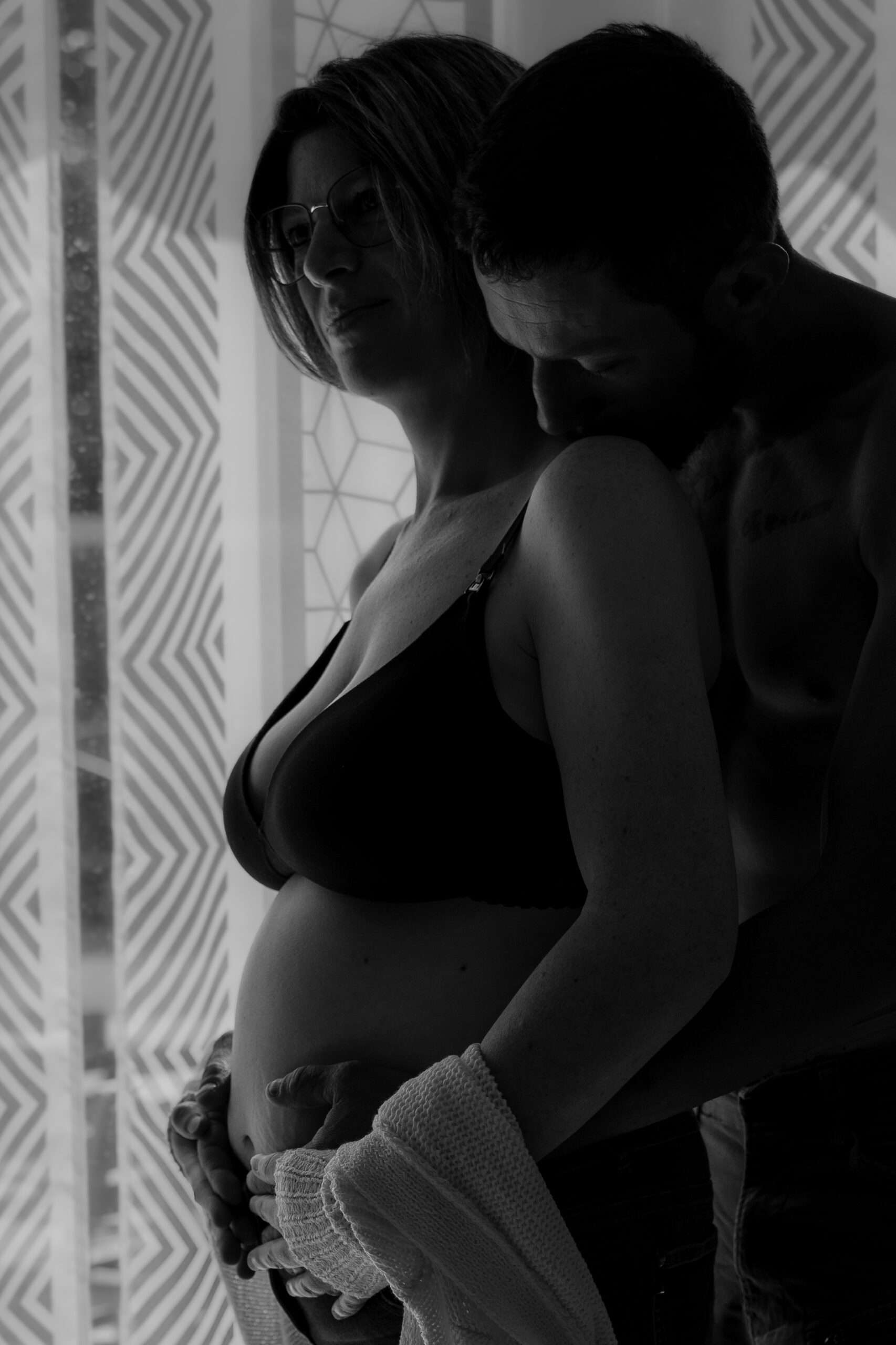 Portrait intime en noir et blanc à domicile d'un couple de futurs parents, le futur père enlaçant dans le dos la future mère, tous les deux les mains posées sur le ventre de la maman