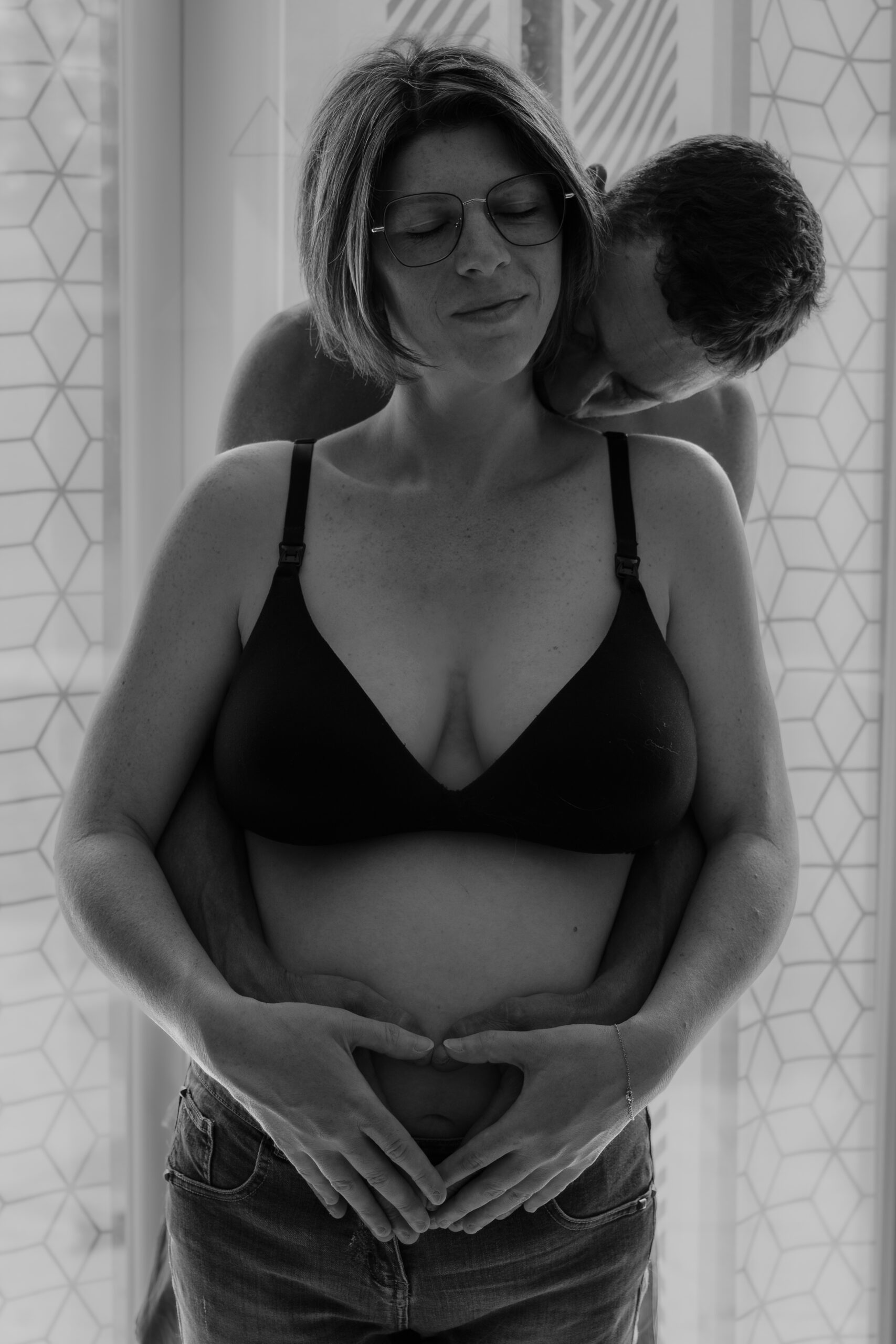 Portrait en noir et blanc de futurs parents de face, le papa enlaçant la maman dans le dos avec leurs mains respective posées sur le ventre de la future mère
