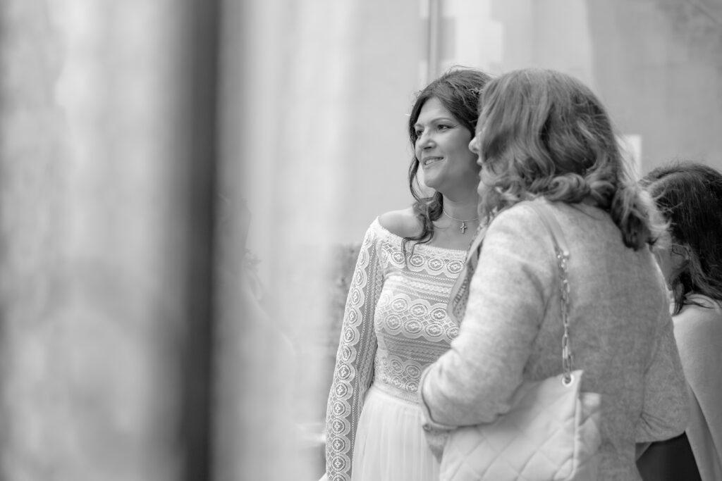 Plan éloigné en noir et blanc de la marié discutant avec une invitée et un premier plan flou encadrant le sujet