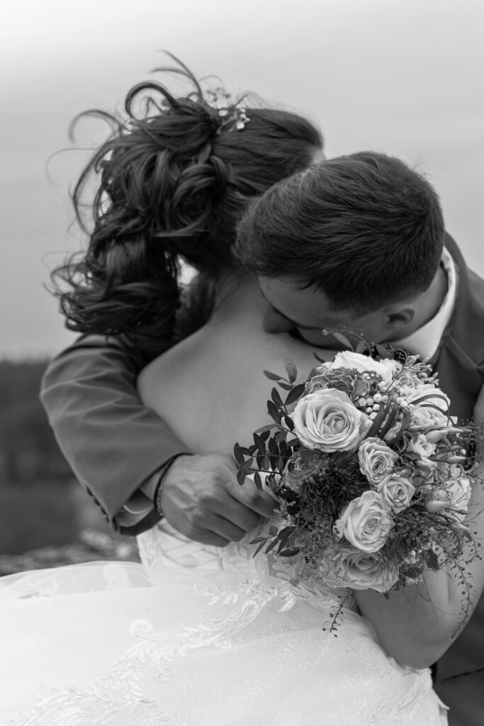Photographie d'un couple lors de leur mariage, le jeune homme enlace la jeune femme tout en tenant son bouquet de fleurs à la main - Manon LORY Photographe