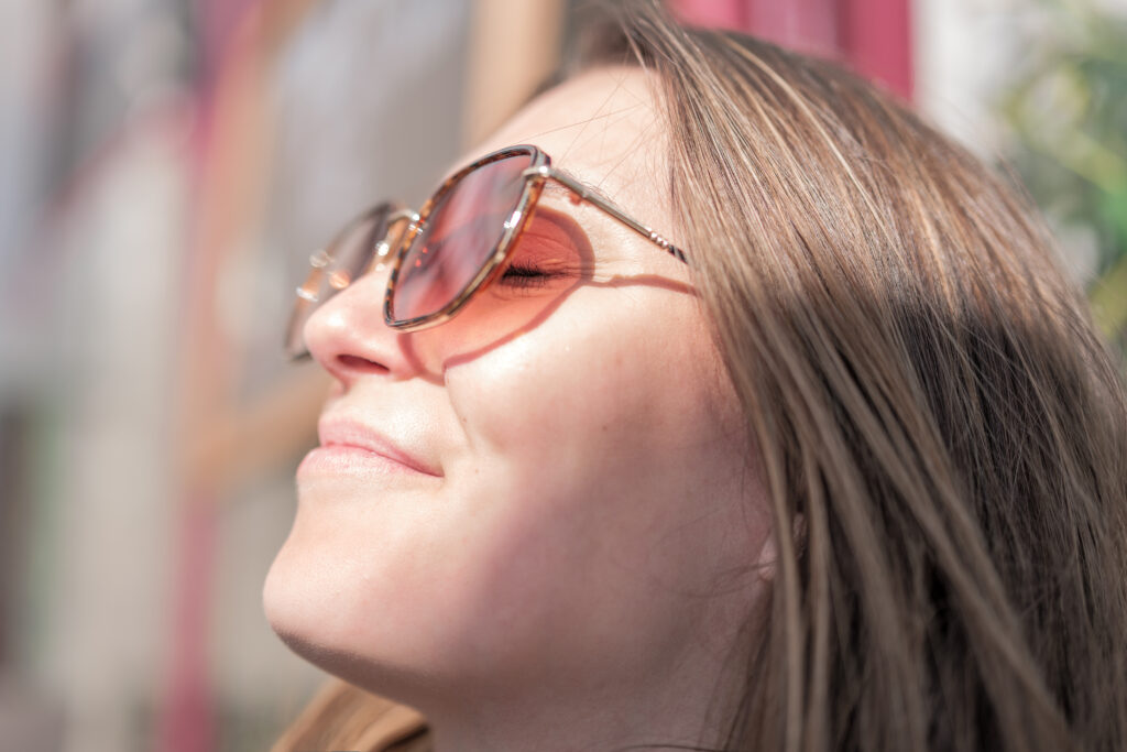Image en plan rapproché d'une femme aux cheveux châtains et portant des lunettes de soleil prenant un bain de soleil en terrasse d'un café