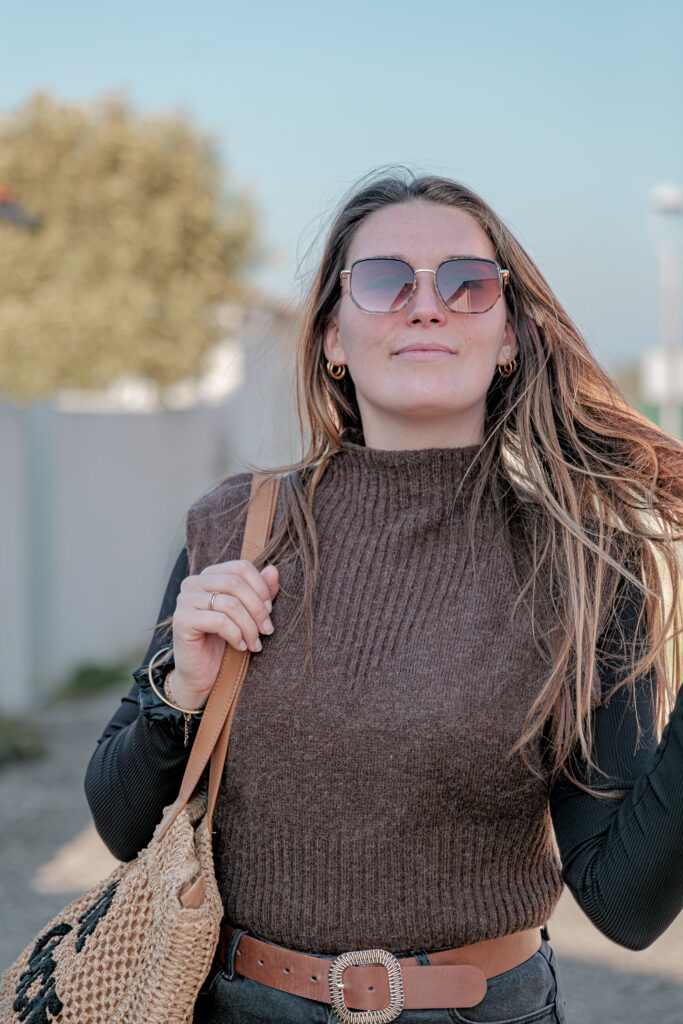 Plan taille d'une femme aux longs cheveux châtains portant des lunettes de soleil un pull sans manches marron et marchant dans une rue de l'île de Ré