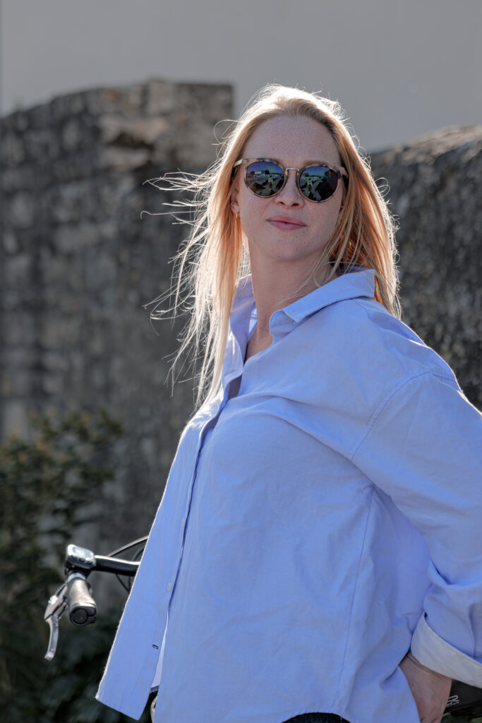 Photographie d'une femme blonde portant des lunettes de soleil et une chemise bleue appuyée sur un vélo
