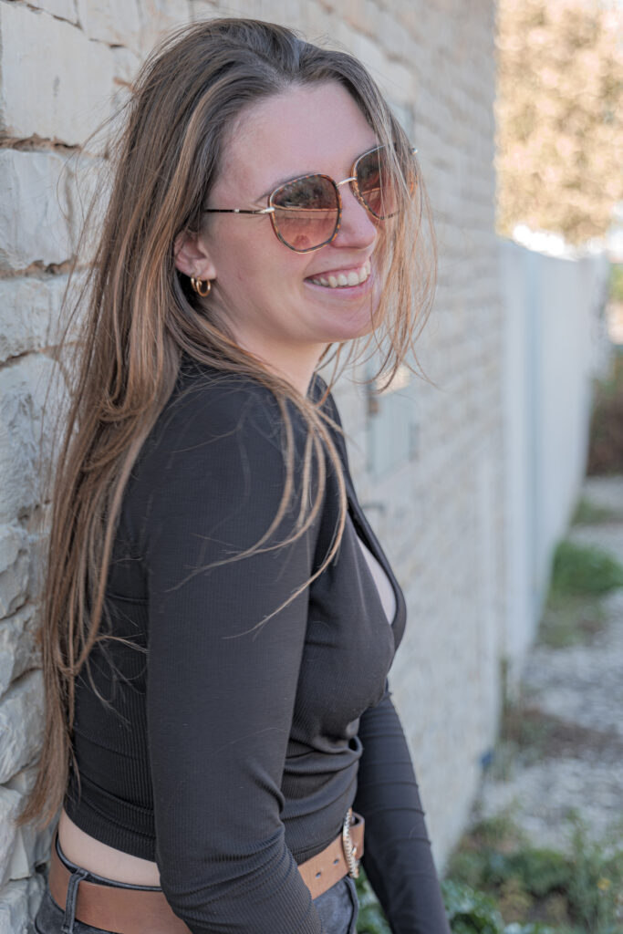 Femme aux long cheveux châtains adossée à un mur en pierre riant avec des lunettes de soleil