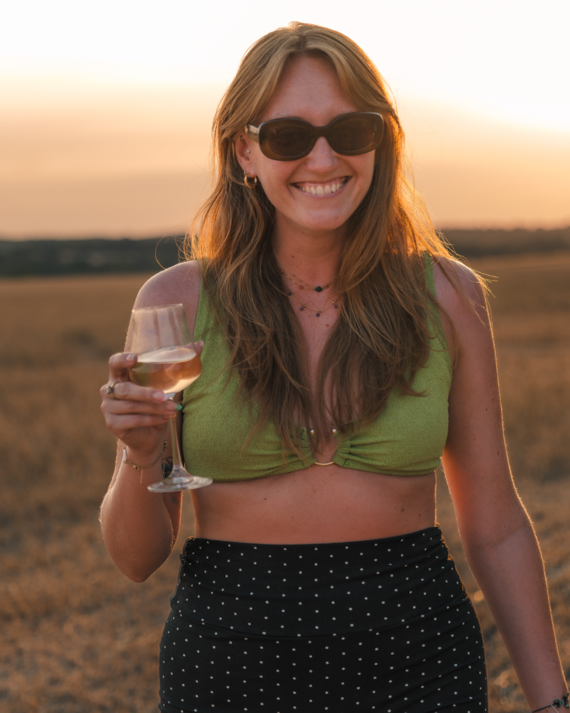 Portrait d'une jeune femme rousse portant des lunettes de soleil avec un verre de vin à la main reflétant le coucher du soleil, réalisé au milieu d'un champ