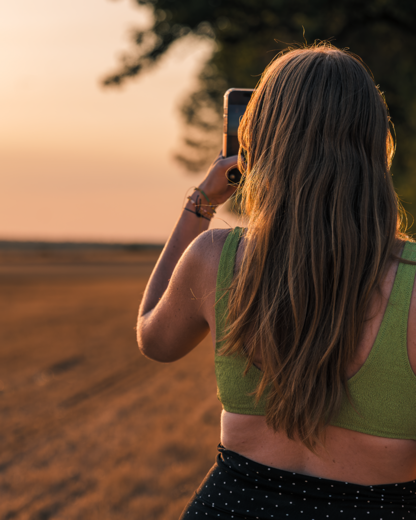 Portrait d'une jeune femme rousse de dos, photographiant avec son téléphone portable le coucher du soleil au milieu d'un champ