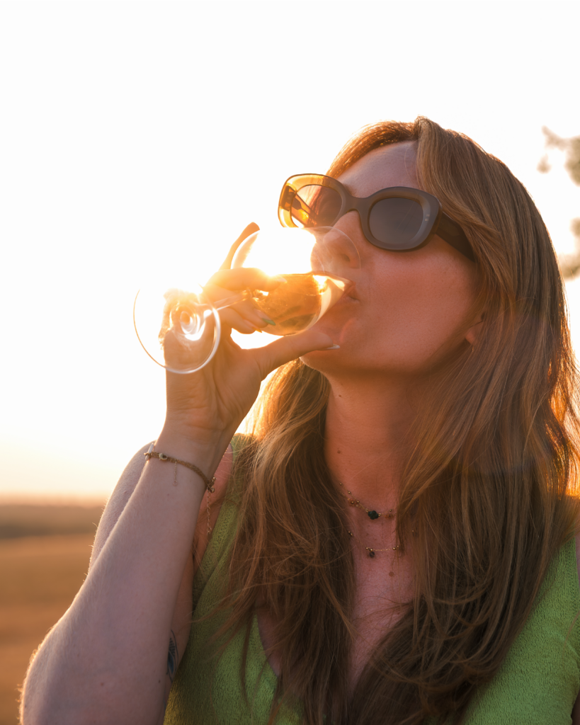 Portrait d'une jeune femme rousse portant des lunettes de soleil avec un verre de vin à la main reflétant le coucher du soleil, réalisé au milieu d'un champ