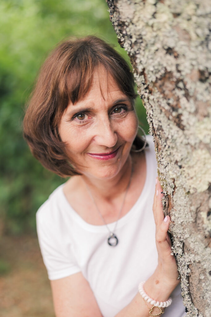 Portrait d'une femme d'âge moyen brune aux yeux noisettes, a demi dissimulée derrière un arbre, photographiée en Creuse avec un arrière plan flou de verdure