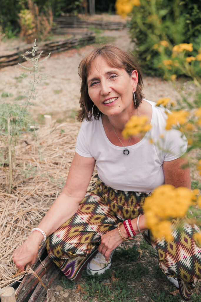Portrait d'une femme d'âge moyen brune aux yeux noisettes, accroupie dans un jardin avec des fleurs jaunes en premier plan