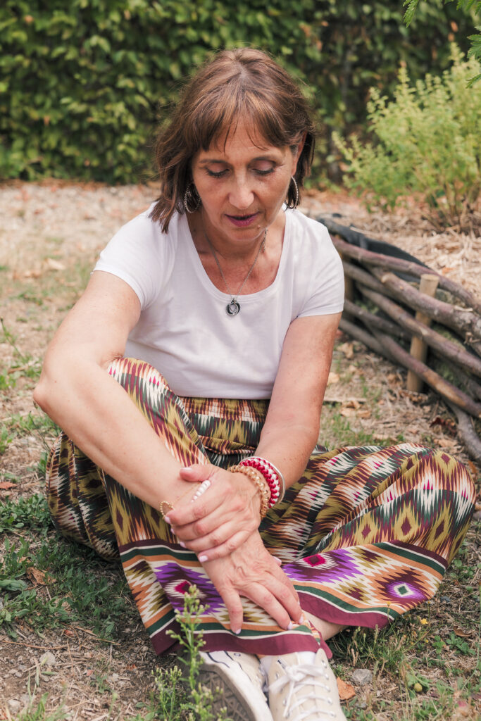 Portrait d'une femme d'âge moyen brune aux yeux noisettes, assise en tailleurs dans un jardin, le regard légèrement orienté vers le bas