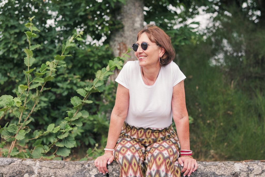 Portrait d'une femme d'âge moyen, assise sur le rebord d'un muret, photographiée en Creuse, portant des lunettes de soleil la tête légèrement tourné vers la gauche avec un arrière plan de verdure