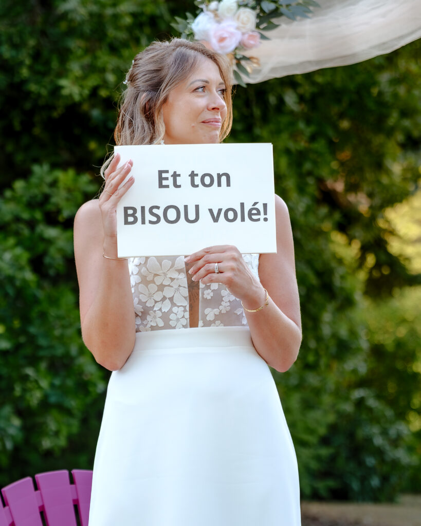 Portrait de la mariée tenant une pancarte avec écrit "Et ton bisou volé" laissant voir un morceau de leur discours silencieux.