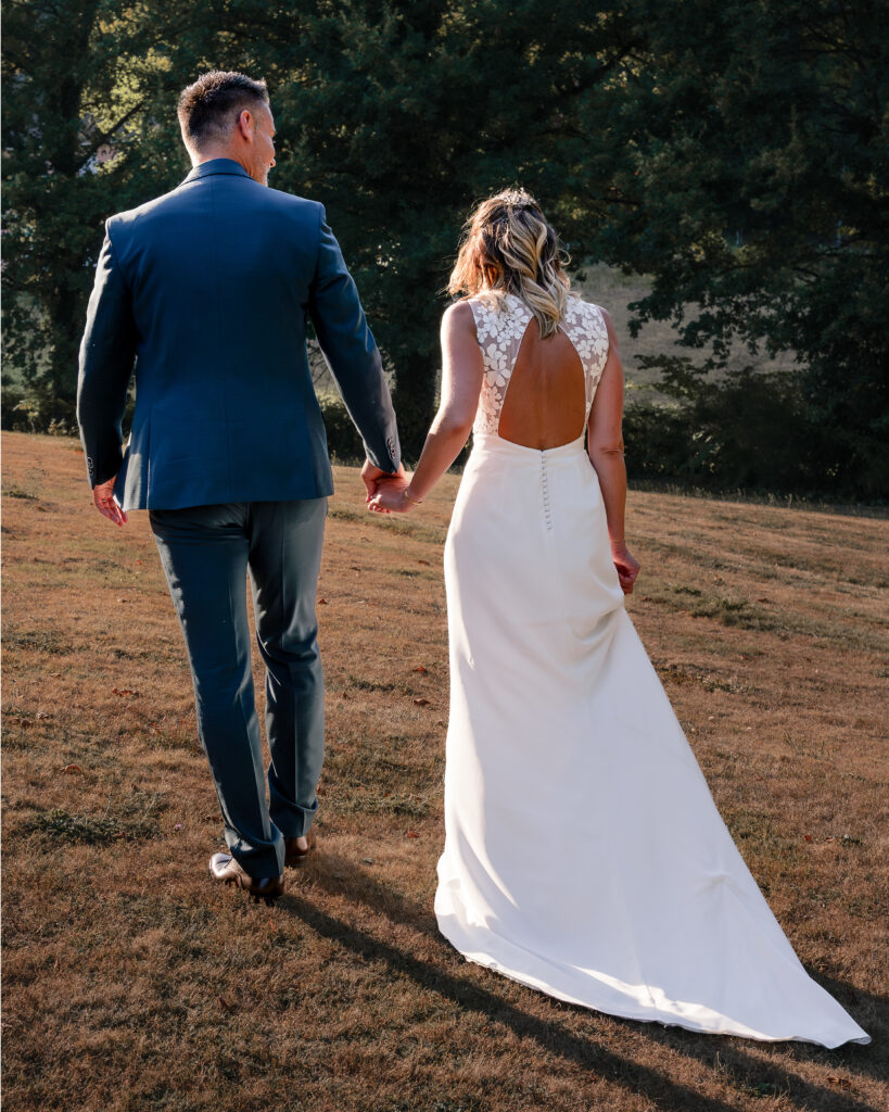 Portrait en pied de dos d'un couple de jeune mariés se tenant la main au coucher du soleil au milieu d'un champ et s'éloignant au loin.