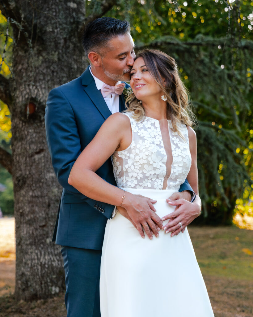 Portrait d'un couple de jeune mariés, le marié enlace la mariée dos à lui en lui embrassant le front