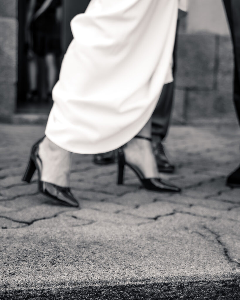 Gros plan à demi flou et en noir et blanc des chaussures à talons de la mariée laissant voir le bitume et donnant un joli contraste avec le bas de la robe blanche.